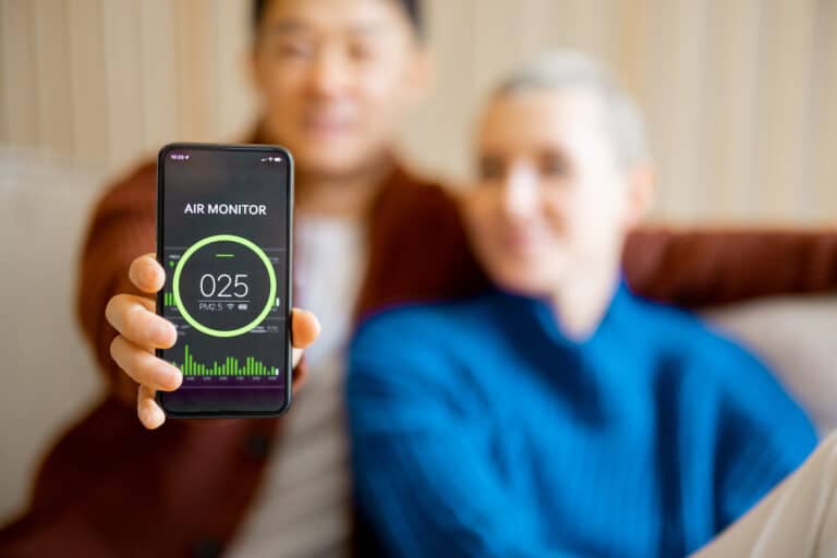 Asian man sitting on sofa with his caucasian wife and showing smartphone with home air condition app. Concept of smart home. Idea of domestic lifestyle. Smiling multiracial couple in modern apartment