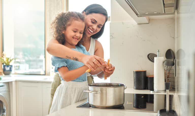 Mother and little daughter cooking together in the kitchen. Mixed race mother and child standing by the stove breaking spaghetti and throwing it in boiling water